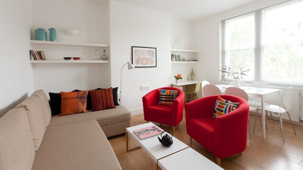Ein modernes Wohnzimmer mit einem beigen Sofa, roten Sesseln, einem weißen Couchtisch und einem Essbereich mit weißem Tisch und Stühlen. Regale mit Büchern und Dekorationselementen sind zu sehen.
