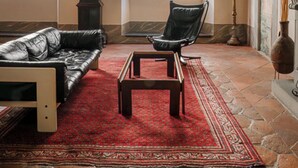 Una habitación con suelo de piedra, un tapete rojo estampado, un sofá de cuero, una mesa de centro de madera, un sillón negro, un reloj alto y una chimenea.