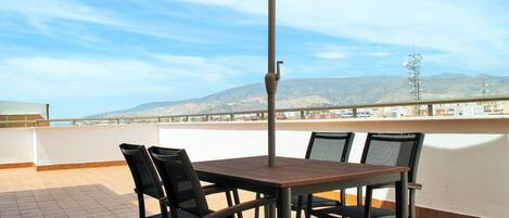 A terrace with a table and chairs, a large umbrella, and a view of mountains and buildings.