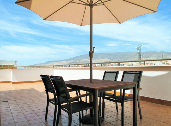 A terrace with a table and chairs, a large umbrella, and a view of mountains and buildings. - PENTHOUSE, TERRACE ON THE HORIZON OF ALMERIA (Almería)