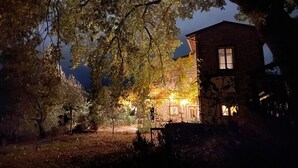 Un bâtiment en pierre doté d’une grande fenêtre et d’une porte, entouré d’arbres et d’un jardin bien éclairé.