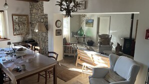 Un salon chaleureux comprenant un coin repas, un mur en pierre, des poutres apparentes, une cheminée et une grande fenêtre laissant entrer la lumière naturelle.