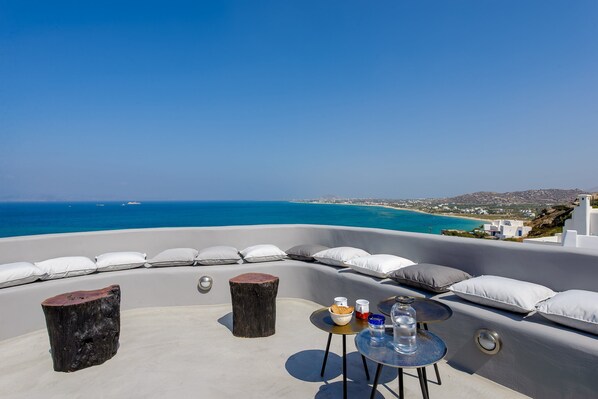 A rooftop terrace with a view of the sea, a round table with a bowl and glasses, and white cushioned seating. - Venti Villa Naxos (Naxos)