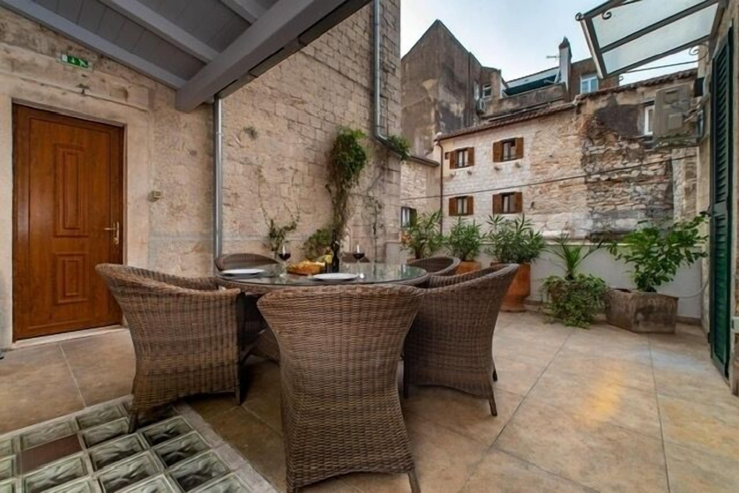 An outdoor seating area with wicker chairs and a glass table, surrounded by stone walls and potted plants.