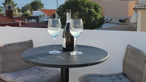 Una mesa redonda con dos vasos y una botella, rodeada de sillas de mimbre con cojines, con vistas a una zona residencial con casas y el mar.