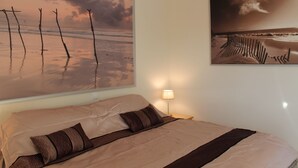 Un dormitorio con cama, almohadas, una mesita de noche con lámpara y dos cuadros decorativos con motivos playeros.