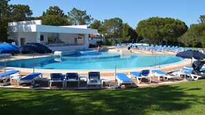 A pool area with lounge chairs and umbrellas, a building with a white facade, and trees in the background. - T1 Duplex in Victory Village - Quinta do Lago (Almancil - ALGARVE)