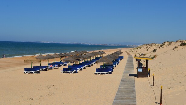 A beach with rows of blue lounge chairs and thatched umbrellas, a wooden boardwalk, and a lifeguard station. - T1 Duplex in Victory Village - Quinta do Lago (Almancil - ALGARVE)
