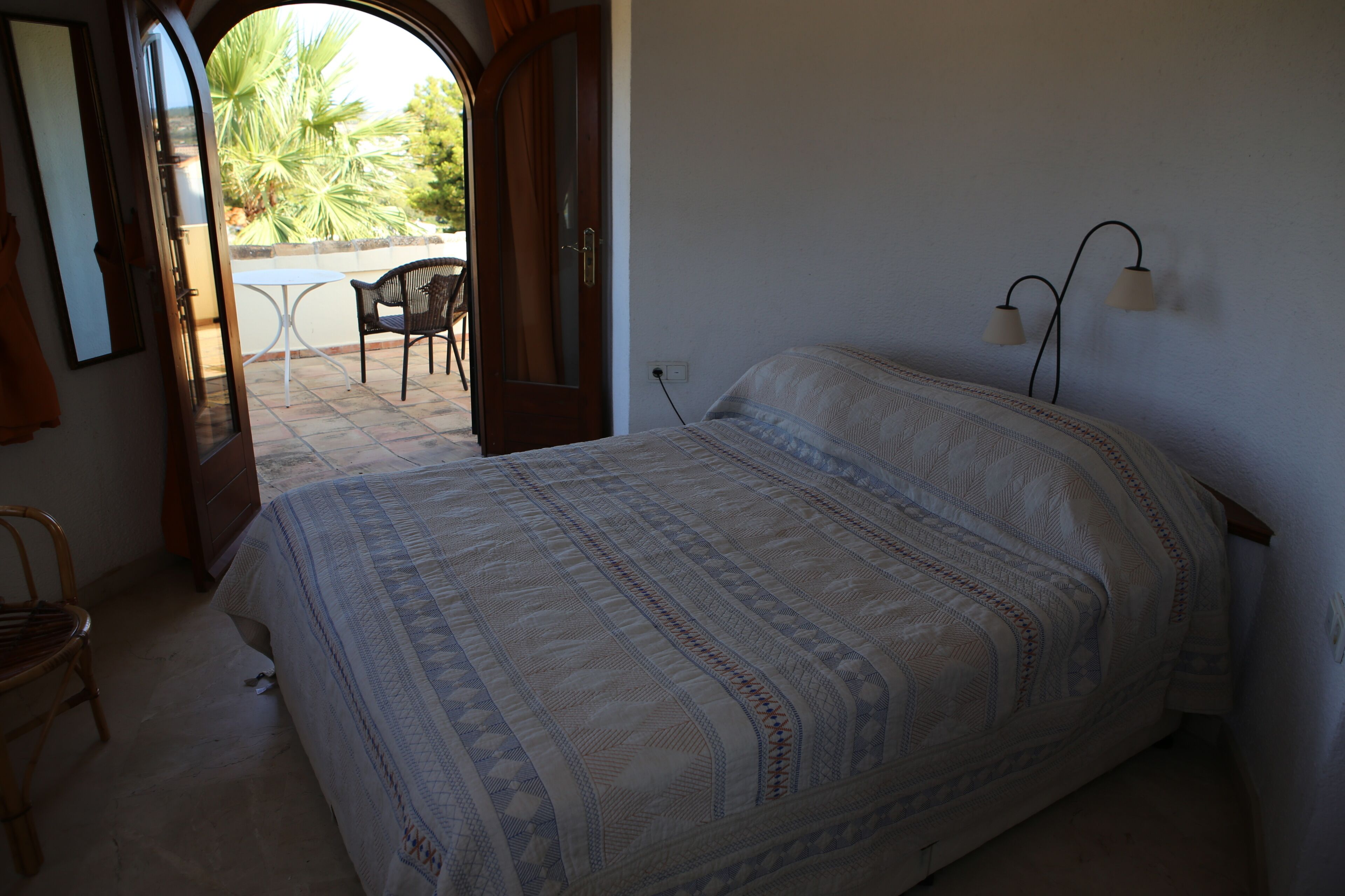 A bedroom with a bed, a chair, and a small table. There is an open door leading to an outdoor area with a table and chairs.