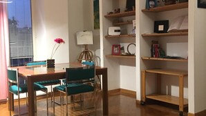Un comedor con una mesa de madera y sillas azul verdosas, una estantería de madera con diversos objetos, y una ventana con persianas.