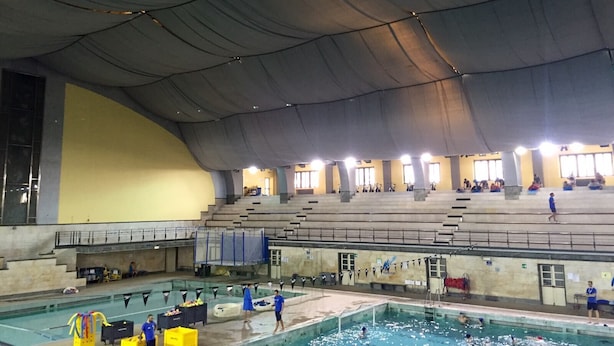 An indoor swimming pool with multiple lanes, a diving platform, and a spectator area with bleachers.