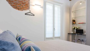 A bedroom with a bed, a hanging light, a brick wall, a window, and a shelf with items.