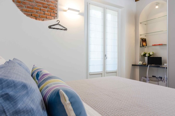 Un dormitorio con cama, una lámpara colgante, pared de ladrillo, ventana y estante con objetos.
