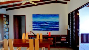 Un comedor con una mesa y sillas de madera, un ventilador de techo, una estantería y un cuadro grande con una escena marina azul.