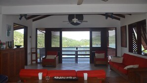Amplia sala de estar con un conjunto de sofá rojo, ventilador de techo y vista a un paisaje verde exuberante a través de ventanas grandes con contraventanas de madera.