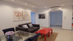 A modern living room with a sofa, a glass coffee table, and a red side table. There is a painting on the wall and a door in the background. - 3 beds/3 baths, 3 min. walking to the Vatican Museums, metro station, FREE WIFI (Roma)