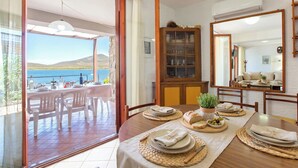 A dining area with a table set for four, a wooden cabinet, and a view of the sea through large glass doors. - ClickSardegna Villa Flavia with seafront terrace in the Porto Conte Nature Park (Alghero)