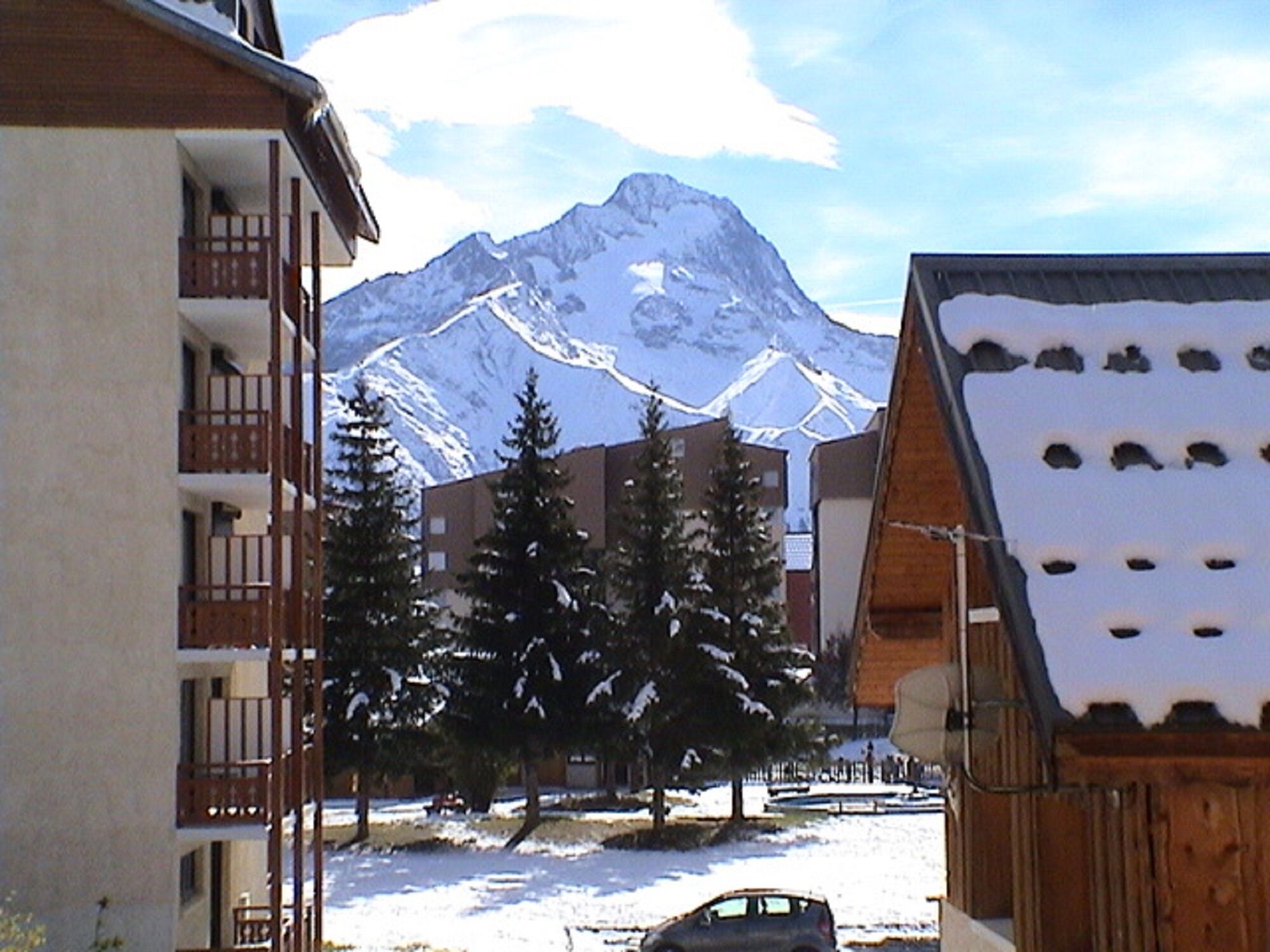 Un edificio con balcón, una montaña nevada al fondo y un coche estacionado al frente.