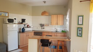 Eine kleine Küche mit Mikrowelle, Kaffeemaschine und Spüle. Holzschränke, ein kleiner Essbereich mit Hockern und ein Fenster mit Vorhängen.