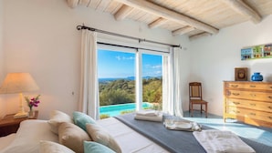 A bedroom with a large bed, a nightstand with a lamp, a chair, a wooden dresser, and a view of the landscape.