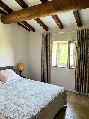 A bedroom with a bed, a nightstand with a lamp, a window with curtains, and wooden beams on the ceiling. - Charming house in the heart of the vineyards (Tarascon)