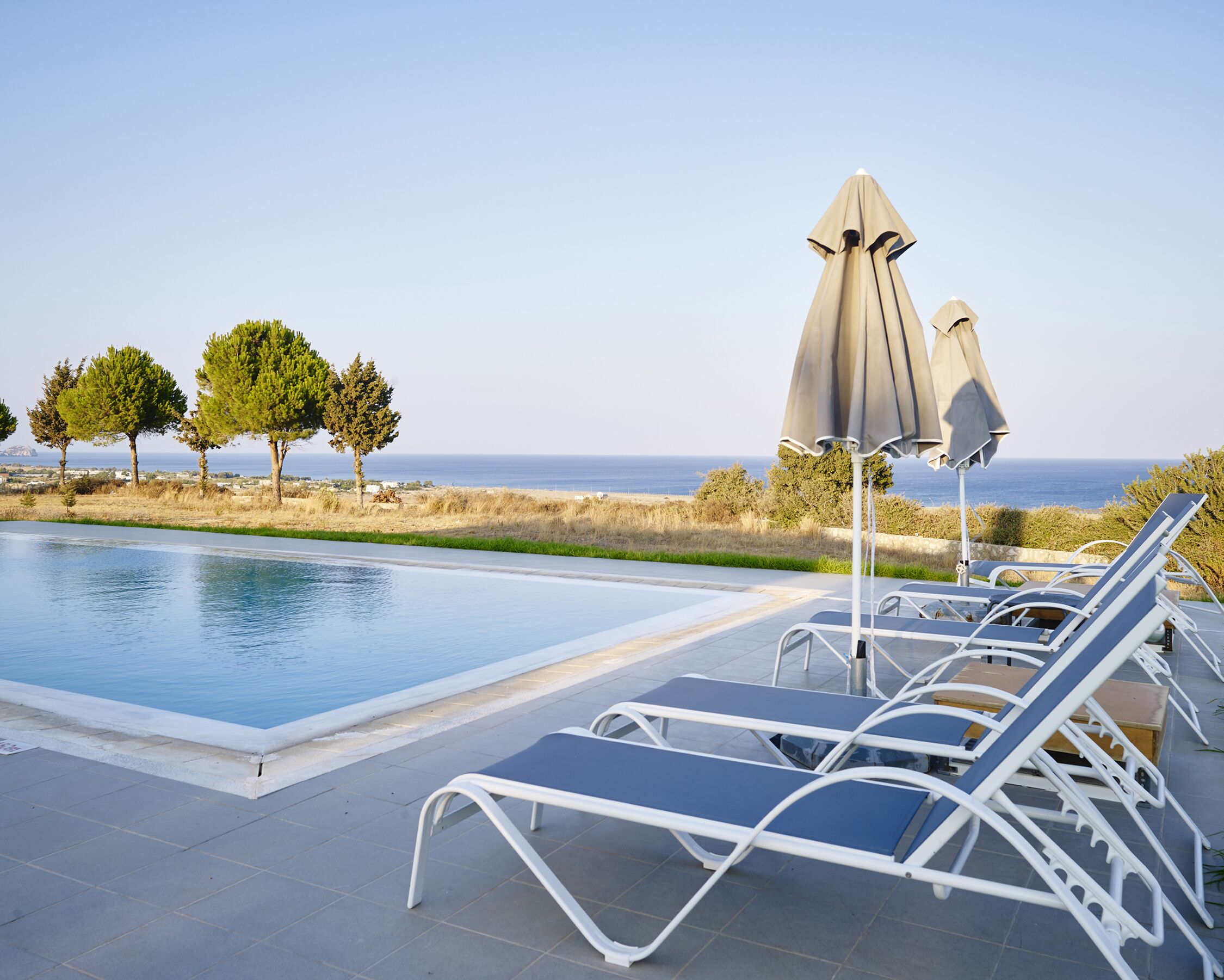 Un espace aménagé au bord de la piscine, avec des chaises longues et des parasols, une vue dégagée sur la mer et quelques arbres en arrière-plan.