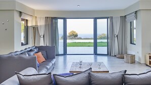 Ein geräumiges Wohnzimmer mit grauem Sofa, einem Couchtisch und Meerblick durch große Schiebeglastüren.