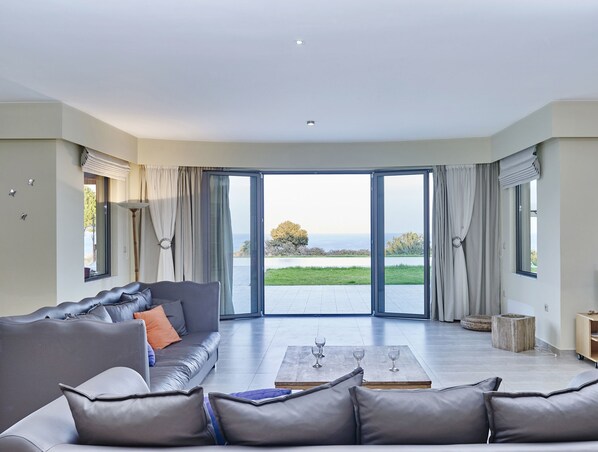 A spacious living room with a grey sofa, a coffee table, and a view of the sea through large sliding glass doors.