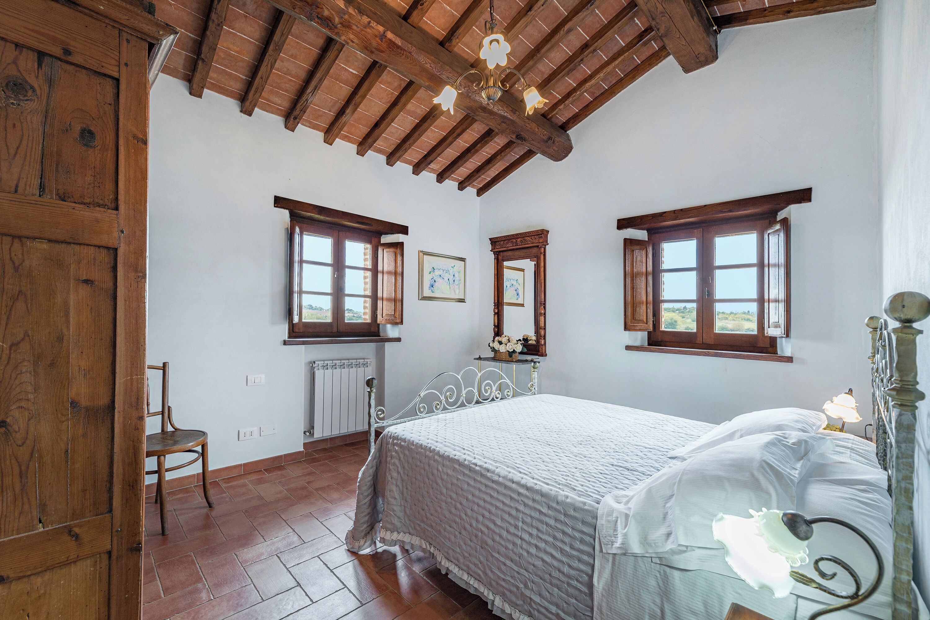 Ein Schlafzimmer mit Holzboden, einem Bett mit weißer Bettwäsche, zwei Fenstern, einem Sessel und einer Heizung.