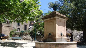 Una plaza pública con una fuente central, rodeada de edificios de piedra y árboles.