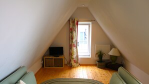 A cozy attic room with a green sofa, a television, a lamp, and a window with a floral curtain. - The Best of Both Worlds - Peaceful Countryside Living with Easy Access to Munich (Reichertshausen)