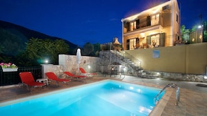 A swimming pool with red lounge chairs, a stone wall, and a building with balconies. - STUNNING TRANQUIL  VILLA WITH PRIVATE POOL CENTRAL LOCATION OF THE ISLAND (Sami)