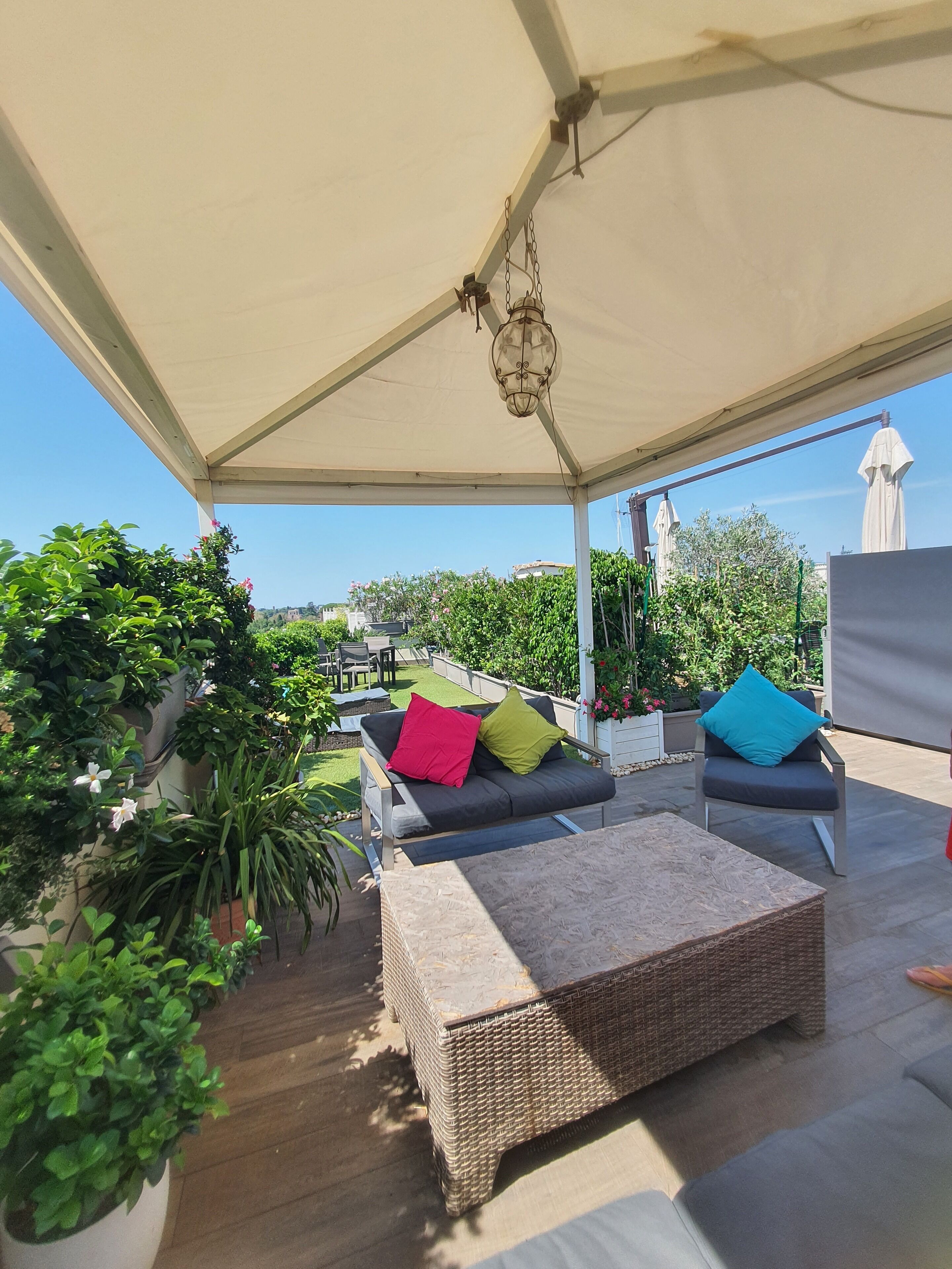 An outdoor patio with a covered seating area, including a sofa and a wicker table, surrounded by potted plants and a view of the cityscape.
