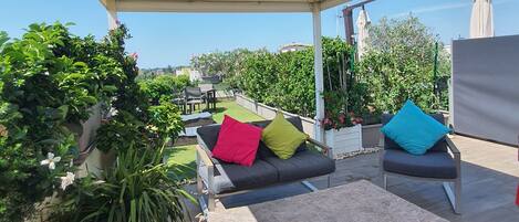 An outdoor patio with a covered seating area, including a sofa and a wicker table, surrounded by potted plants and a view of the cityscape.