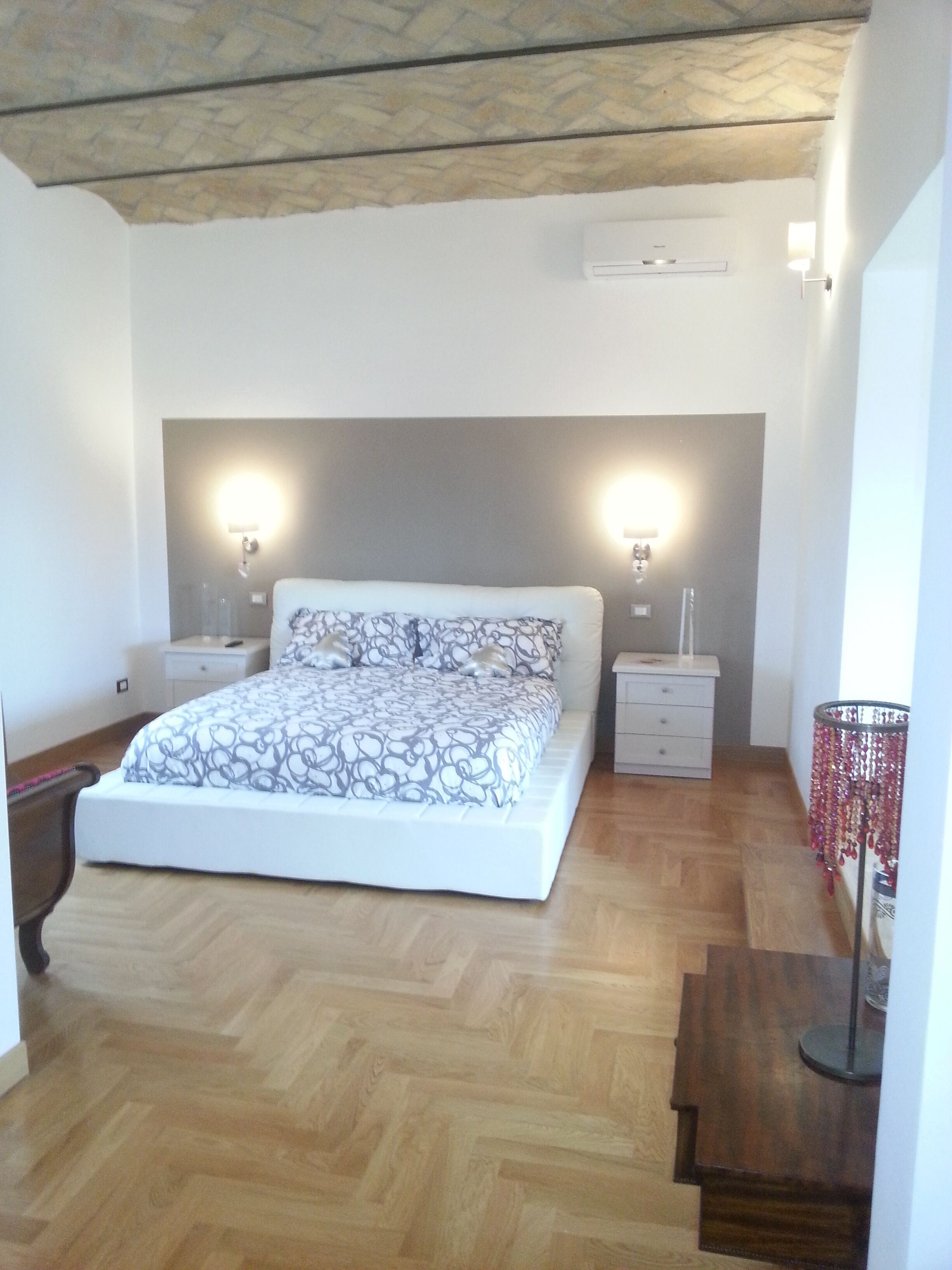 A bedroom with a large bed, two bedside tables, a nightstand, a lamp, and a wooden floor.