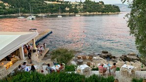 Ein Restaurant am Wasser mit Außenbestuhlung und Meerblick, umgeben von viel Grün und Felsen.