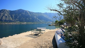 A bench by a lake with mountains in the background. - The Stone House, Prcanj 165 - Seafront Apartment. (Kotor)