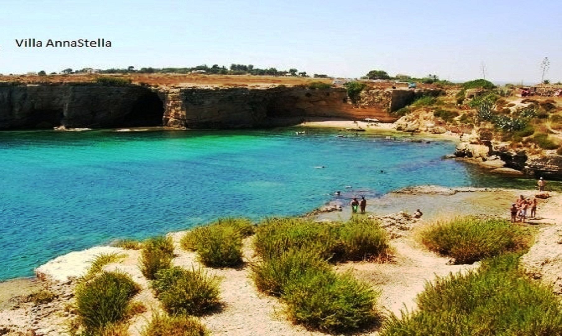 A coastal landscape with clear blue water, rocky cliffs, and sparse vegetation.