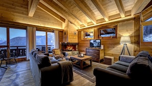 L’intérieur chaleureux d’un chalet en bois, avec une cheminée, des sièges confortables et de grandes fenêtres offrant une vue imprenable sur les montagnes.