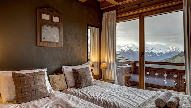 Une chambre avec un grand lit, une tête de lit en bois, une plaque nominative fixée au mur, et une vue sur des montagnes enneigées par une porte vitrée.