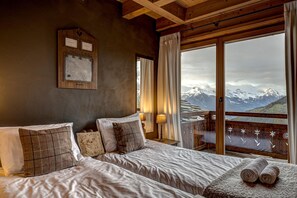 Une chambre avec un grand lit, une tête de lit en bois, une plaque nominative fixée au mur, et une vue sur des montagnes enneigées par une porte vitrée.