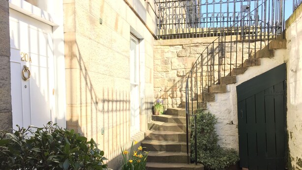 Un estrecho patio con una escalera de piedra que sube a un segundo piso. Se observan macetas con plantas y una puerta verde a la derecha.