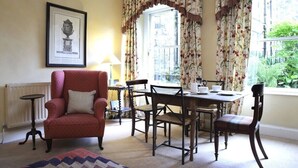 Una habitación con un sillón rojo, una mesa con sillas, un cuadro en la pared y una ventana con cortinas florales.