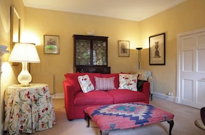 A cozy living room with a red sofa, a patterned ottoman, a cabinet with glass doors, and a lamp on a side table. - Elegant 2 bedroom garden apartment in Stockbridge (Edinburgh)