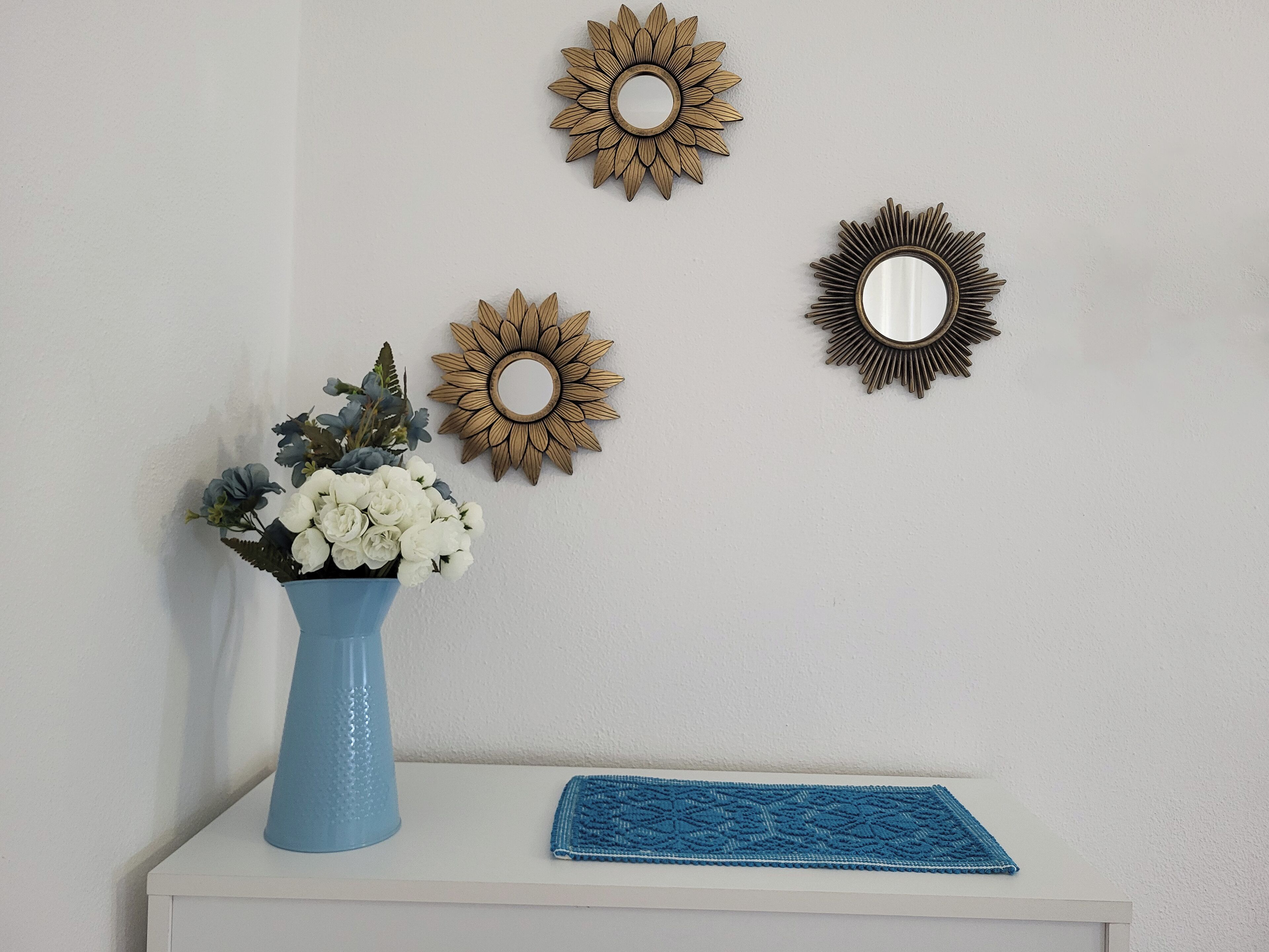 Un mur blanc orné de trois miroirs en forme de soleil et d’un vase bleu contenant des fleurs blanches, le tout posé sur une étagère blanche.