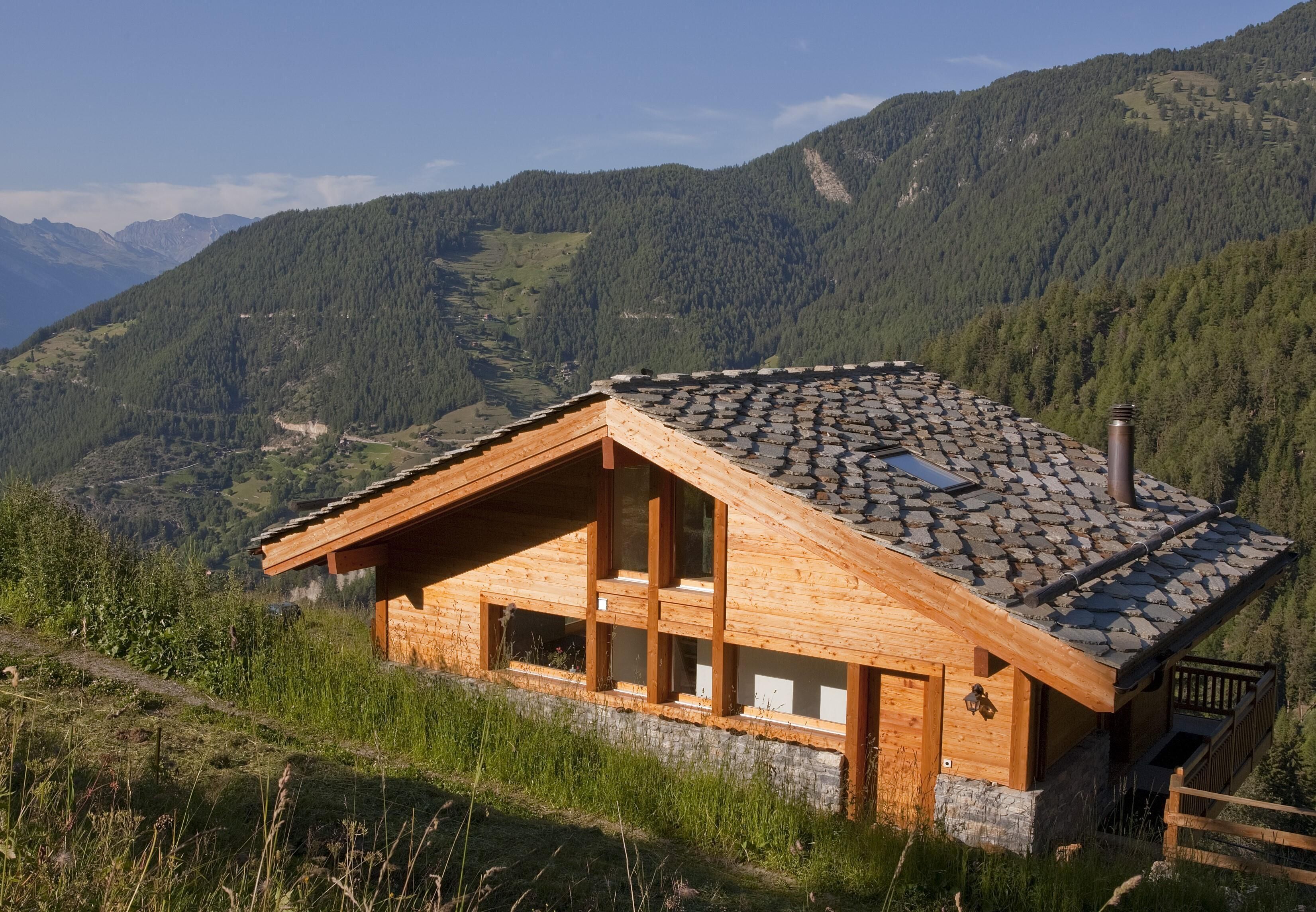 A wooden cabin with a sloped roof, large windows, and a stone foundation, surrounded by forested mountains.