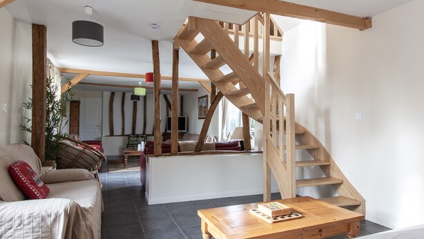 Een ruime woonkamer met een houten trap, een bank, een salontafel en een zicht op de keuken in de achtergrond.
