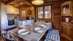 Interior de una cabaña de madera con una mesa de comedor preparada para seis personas, electrodomésticos de cocina y un ventanal grande.