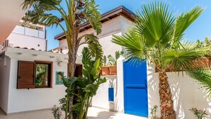 A courtyard with a white building, a blue door, and various tropical plants. - PRIVATE POOL 3 BEDROOMS 3 BATHROOMS 150 MT FROM THE SEA WI FI FREE , CAR RENTAL (CASTELLAMMARE DEL GOLFO)