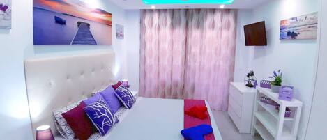 A modern bedroom with a large bed, a headboard, a bedside table, a TV, and a window with curtains.